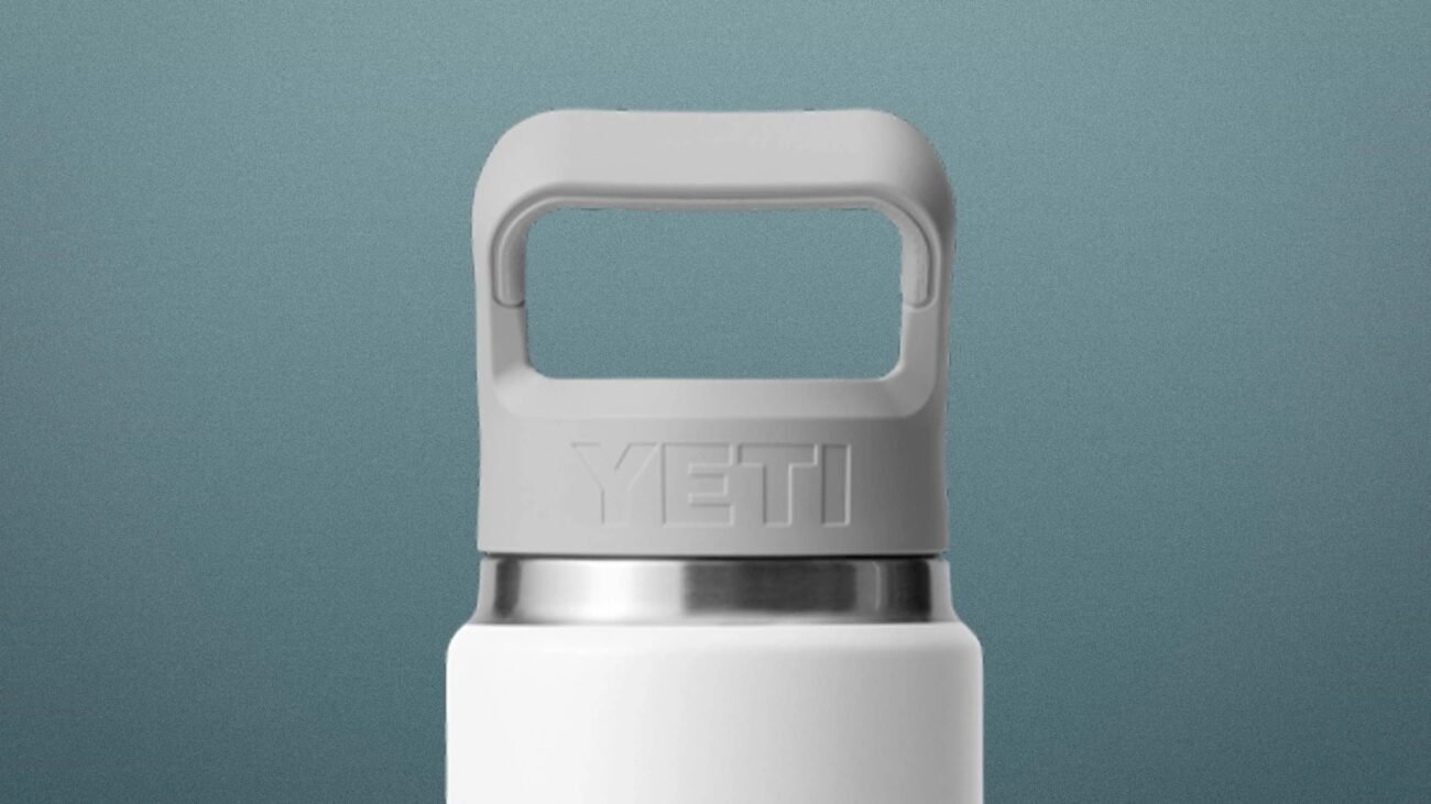 White YETI bottle with a gray handle cap and stainless steel rim against a blue-gray background.